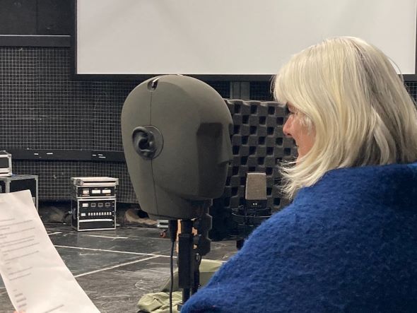A oman with white hair and a blue scarf is facing a microphone shaped like a head.