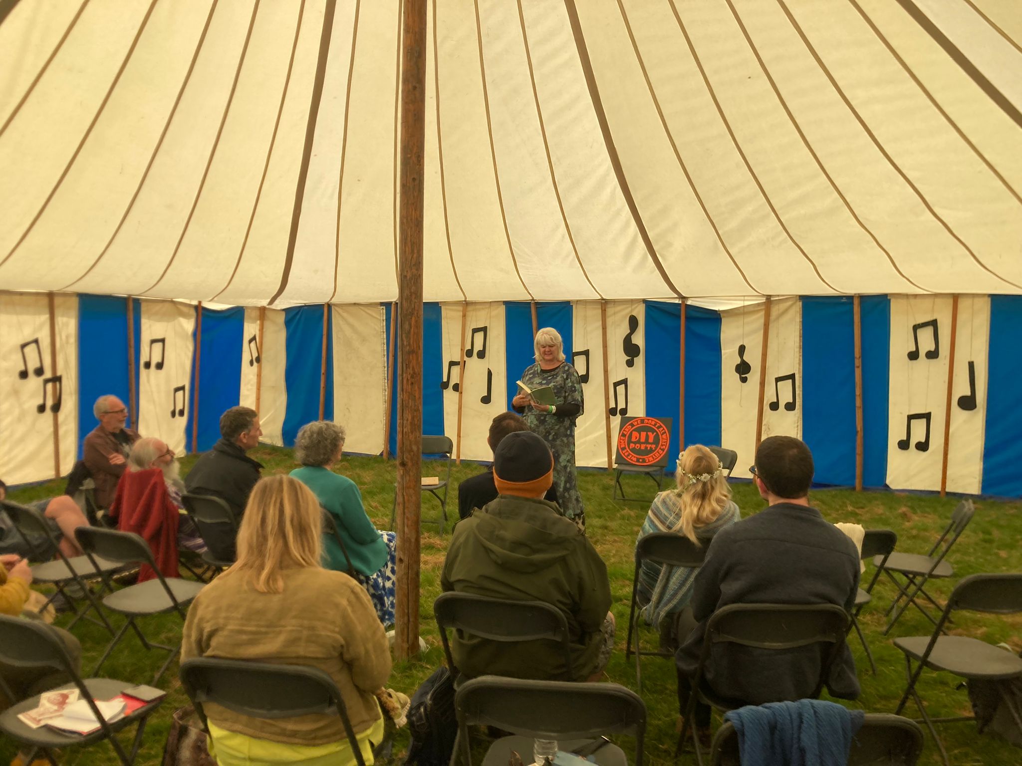 Audience sits facing away watching Lytisha perform. Behind her are blue and white panelled walls of the tent. In the white sections cut out music notes decorate the space.