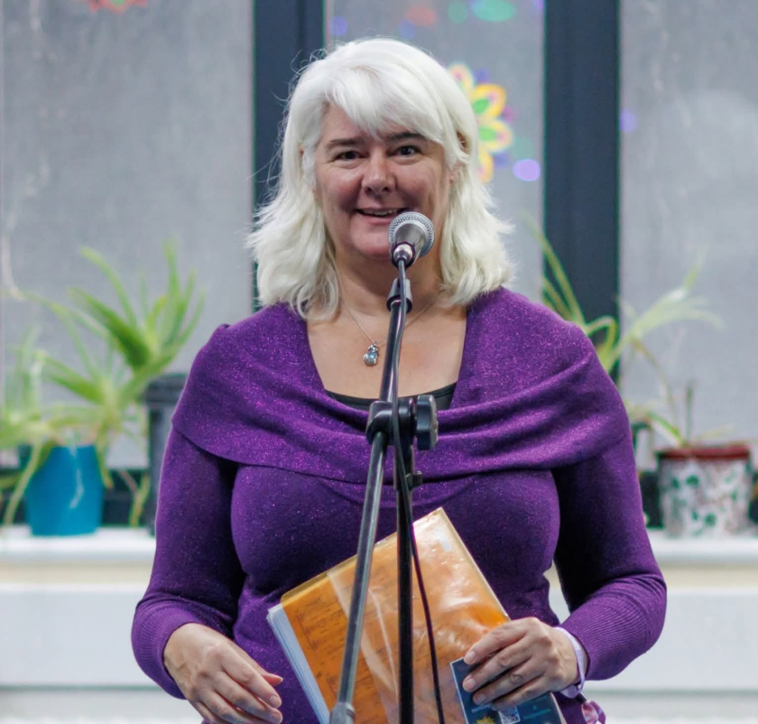 White woman in 50's with white hair, purple top, holding folder of opems, standing behind microphone.